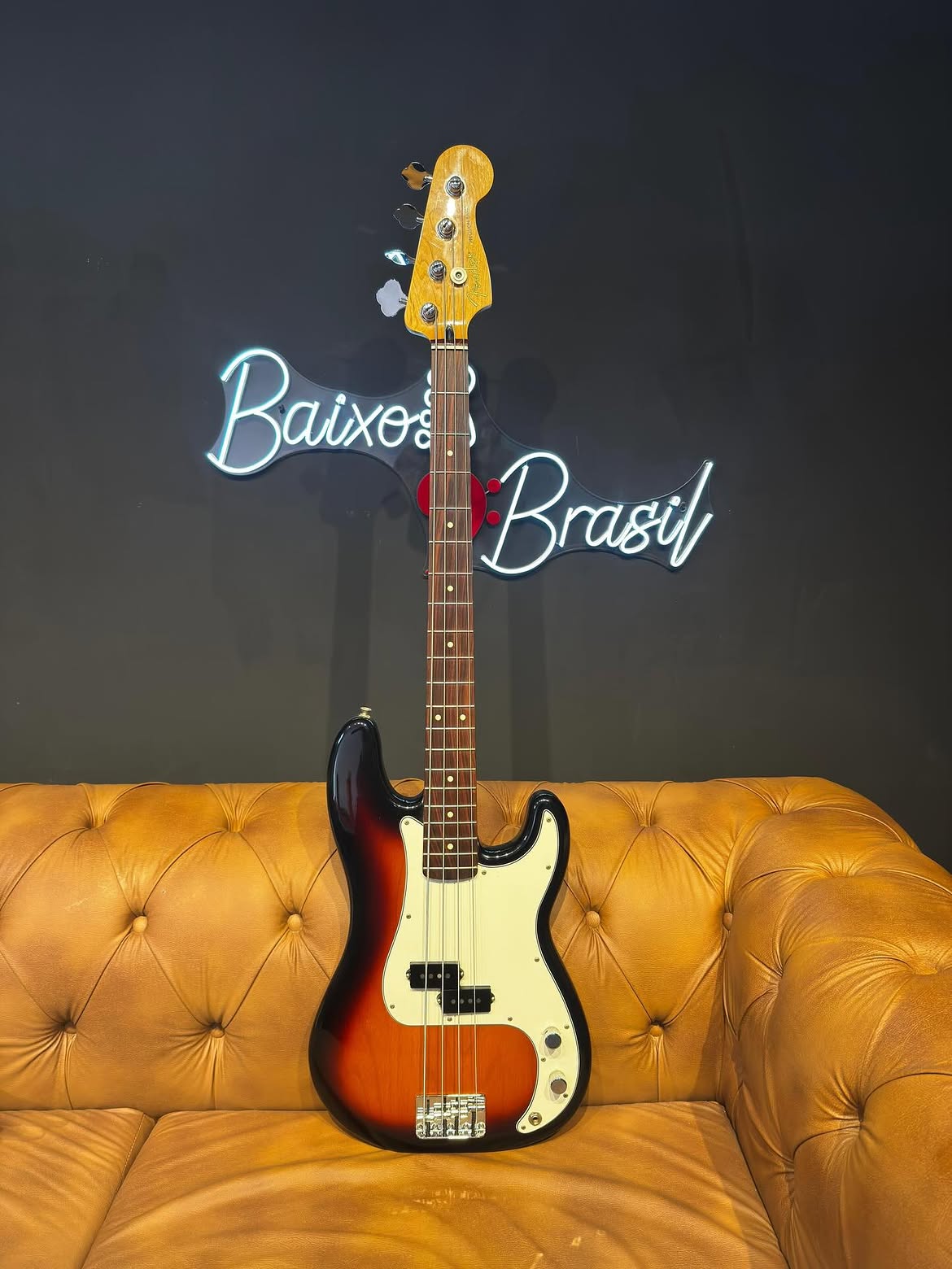 Fender Precision Bass sunburst sobre um sofá de couro, em frente ao letreiro neon da Baixo Brasil